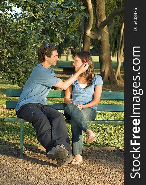 Happy couple on a park bench smile at each other as he places a red flower behind her ear. Vertically framed photograph. Happy couple on a park bench smile at each other as he places a red flower behind her ear. Vertically framed photograph.