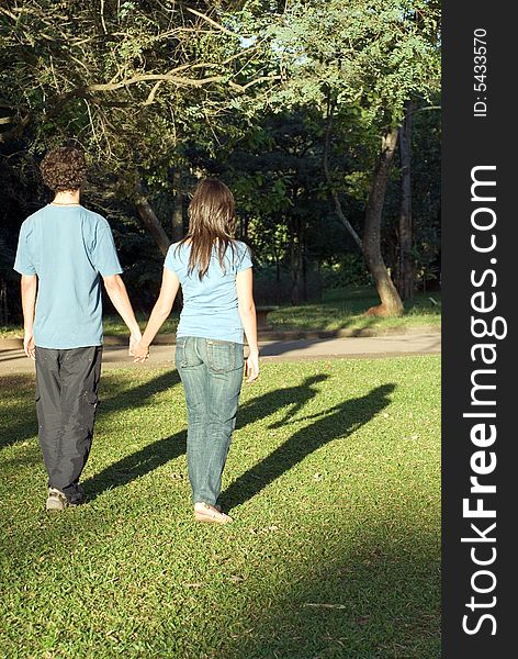 Young couple walking through a park holding hands on a sunny day. They are facing away and you can see their shadows. Vertically framed photograph. Young couple walking through a park holding hands on a sunny day. They are facing away and you can see their shadows. Vertically framed photograph