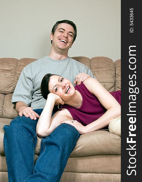 Smiling, happy couple sitting on a couch. They are laughing and he has his arm around her as she lays across his lap. Vertically framed photograph. Smiling, happy couple sitting on a couch. They are laughing and he has his arm around her as she lays across his lap. Vertically framed photograph
