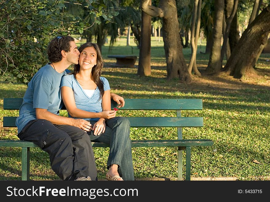 A man kisses a woman's cheek as they sit on a bench. She smiles and looks away and he has his arm around her. Horizontally framed photograph. A man kisses a woman's cheek as they sit on a bench. She smiles and looks away and he has his arm around her. Horizontally framed photograph.