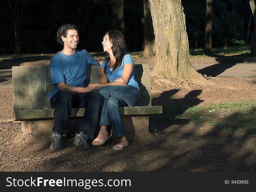 Happy, laughing couple holding hands as they sit on a stone bench in a shaded park. Horizontally framed photograph. Happy, laughing couple holding hands as they sit on a stone bench in a shaded park. Horizontally framed photograph