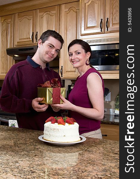 Happy couple in a kitchen smiling near a cake. Both are holding a present while smiling at the camera. Vertically framed photograph. Happy couple in a kitchen smiling near a cake. Both are holding a present while smiling at the camera. Vertically framed photograph.