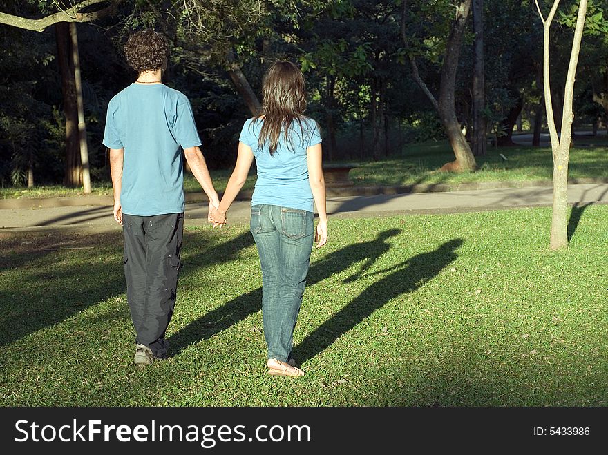 Young couple walking through a park holding hands on a sunny day. They are facing away and you can see their shadows. Horizontally framed photograph. Young couple walking through a park holding hands on a sunny day. They are facing away and you can see their shadows. Horizontally framed photograph