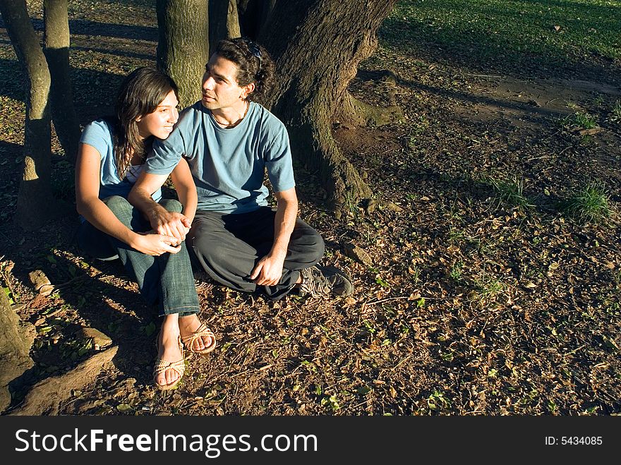 Happy couple sitting on the ground under the trees. She is resting her head on his shoulder as he looks away. Horizontally framed photograph. Happy couple sitting on the ground under the trees. She is resting her head on his shoulder as he looks away. Horizontally framed photograph.