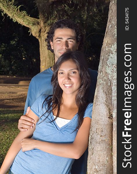 Portrait of a happy couple leaning against a tree in a park. They are smiling as he holds her arm. Vertically framed photograph. Portrait of a happy couple leaning against a tree in a park. They are smiling as he holds her arm. Vertically framed photograph.