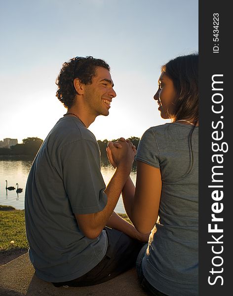 A smiling couple holding hands and gazing at each other front of a beautiful pond with swans swimming in it. Vertically framed photograph. A smiling couple holding hands and gazing at each other front of a beautiful pond with swans swimming in it. Vertically framed photograph.