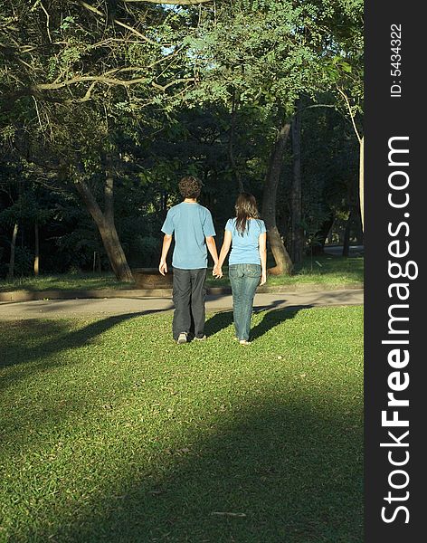 Young couple walking through a park holding hands on a sunny day. They are facing away and you can see their shadows. Vertically framed photograph. Young couple walking through a park holding hands on a sunny day. They are facing away and you can see their shadows. Vertically framed photograph