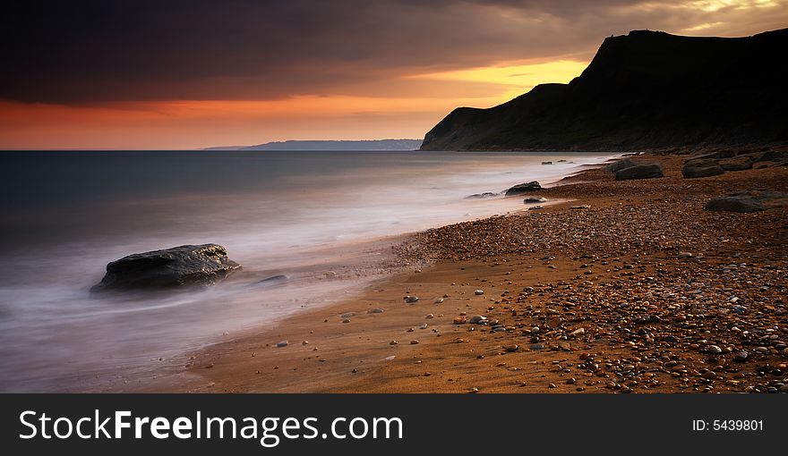 The Sun setting behind the picturesque cliffs of Eype beach in Dorset. A long exposure has resulted in soft movement in the waves as they crash on the pebble and sand beach. The Sun setting behind the picturesque cliffs of Eype beach in Dorset. A long exposure has resulted in soft movement in the waves as they crash on the pebble and sand beach