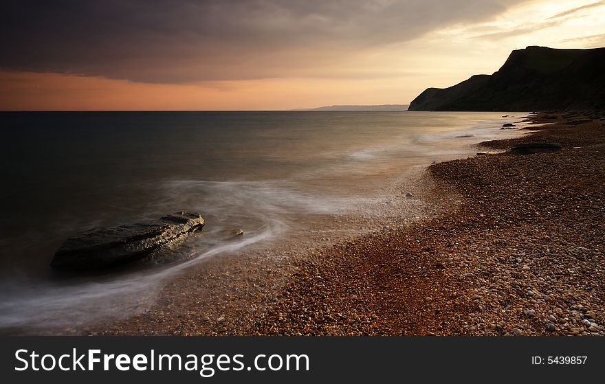 The Sun setting behind the picturesque cliffs of Eype beach in Dorset. A long exposure has resulted in soft movement in the waves as they crash on the pebble and sand beach. The Sun setting behind the picturesque cliffs of Eype beach in Dorset. A long exposure has resulted in soft movement in the waves as they crash on the pebble and sand beach