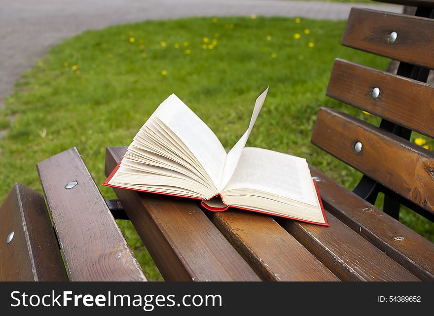 Open book in the red cover lies on a park bench on sunny spring day