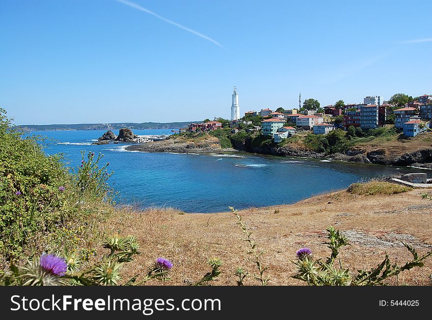 A village with lighthouse at a cape with rocky shore breaking waves viewed form behind lilac wildflowers, under blue sky with a white streak of contrail. Fener village at the tip of Black Sea and Bosphorus.