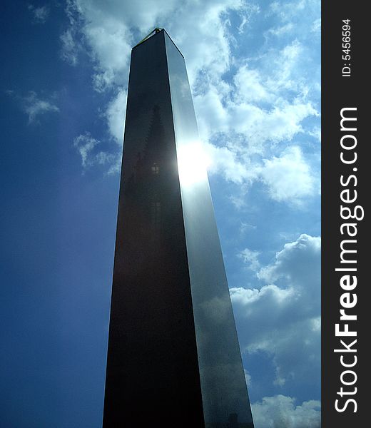 An obelisk on sky with a cathedral and sun reflection.