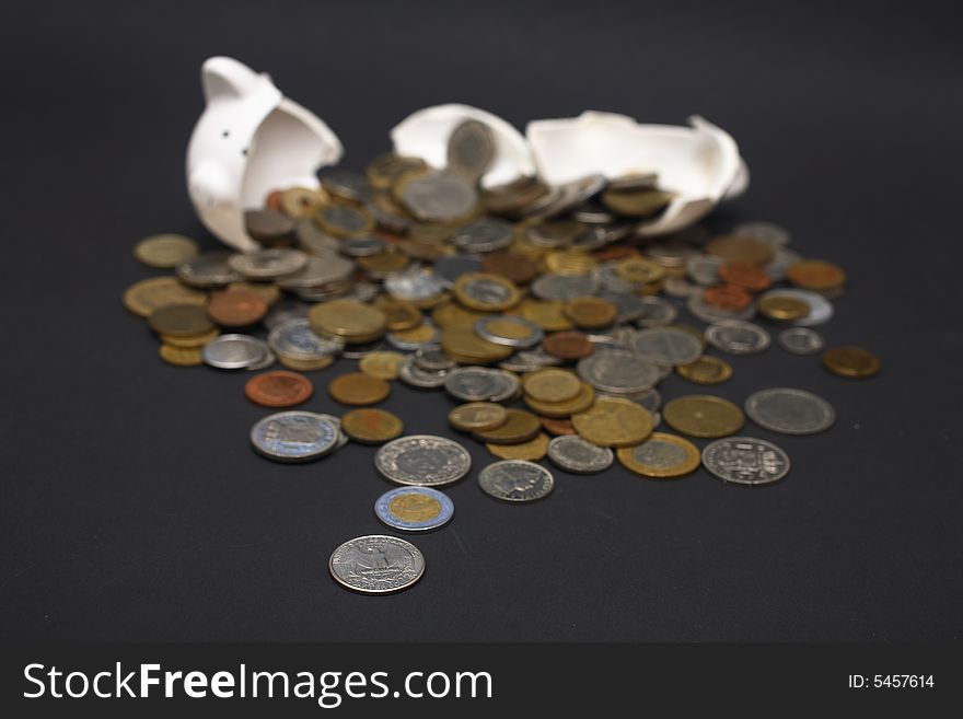 A broken piggy bank isolated on a dark background with loads of coins from around the world.