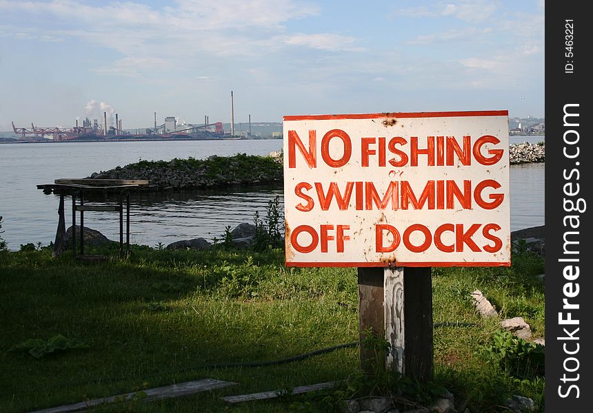 A sign warns of no swimming or fishing off dock, with industry in the background. A sign warns of no swimming or fishing off dock, with industry in the background.