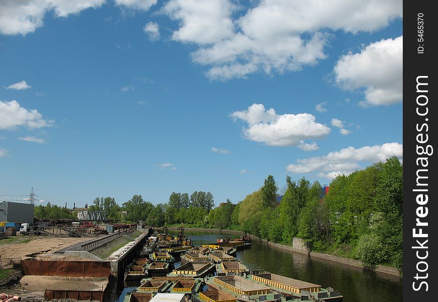 The barges on the canal. The industrial water landscape. The blue sky with overcast. Trees on the bank. Spring.