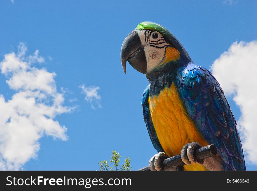 A giant stature of a blue and yellow macaw parrot on a perch with a blue sky and cloud background. Copy space to the left. A giant stature of a blue and yellow macaw parrot on a perch with a blue sky and cloud background. Copy space to the left.