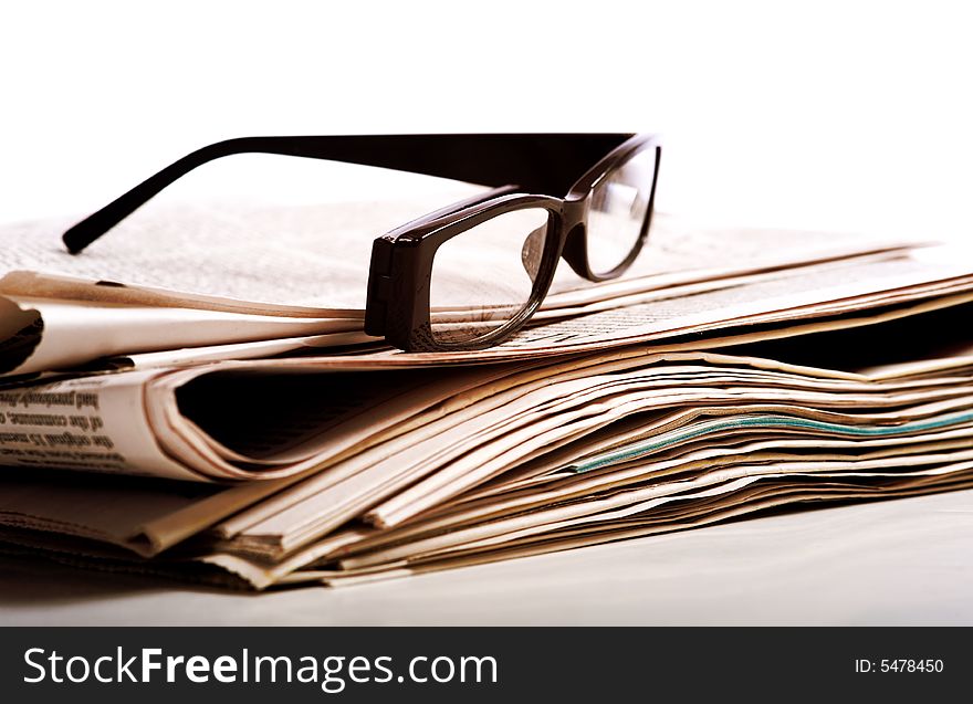 A pair of black reading glasses on top of a stack of newspapers on a white background. A pair of black reading glasses on top of a stack of newspapers on a white background