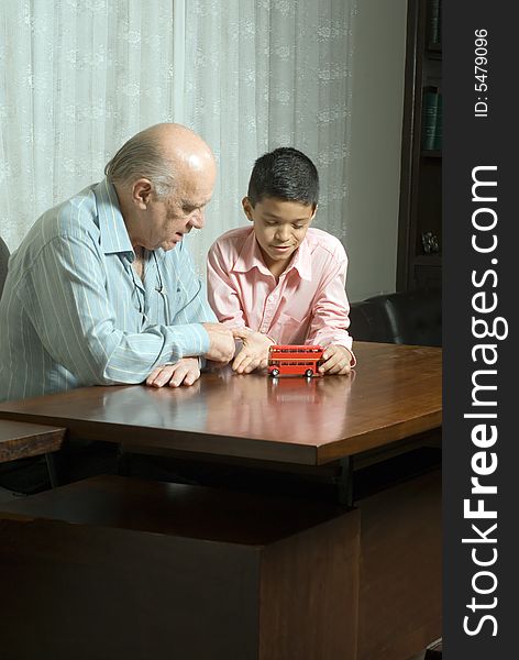 Grandfather and grandson sitting at the table with a toy bus. Grandson is playing with the bus as grandfather watches. This is a horizontally framed photo. Grandfather and grandson sitting at the table with a toy bus. Grandson is playing with the bus as grandfather watches. This is a horizontally framed photo.