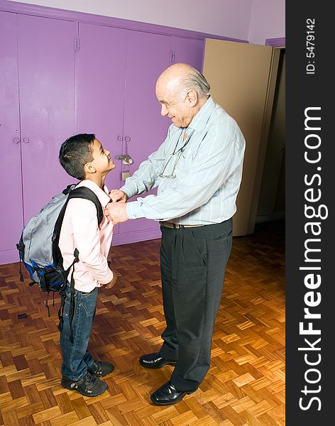 Grandfather is buttoning grandsons shirt as he is leaving for school. Grandson stands there patiently with his backpack smiling as grandfather helps him. This is a vertically framed photo. Grandfather is buttoning grandsons shirt as he is leaving for school. Grandson stands there patiently with his backpack smiling as grandfather helps him. This is a vertically framed photo.