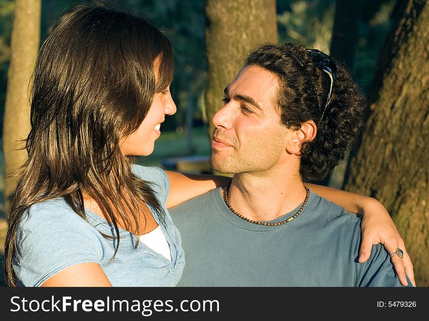 Couple staring at each other. She is smiling at him and he looks serious. Trees in the background. Horizontally framed photograph. Couple staring at each other. She is smiling at him and he looks serious. Trees in the background. Horizontally framed photograph.