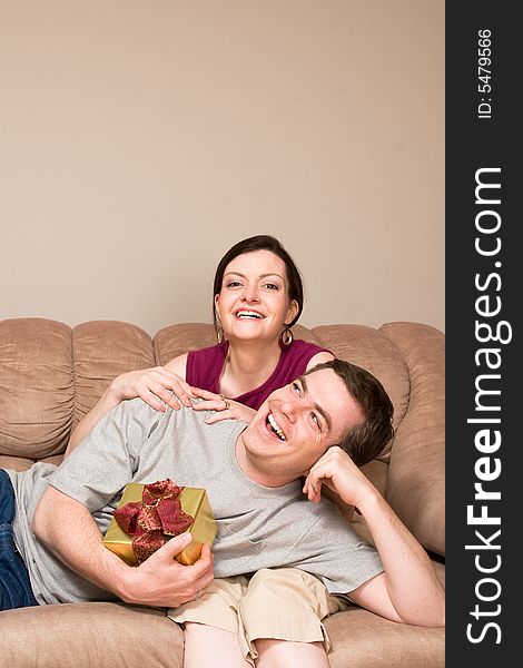 A smiling man holding a gift box lying on a smiling woman's lap on sofa.  Vertically framed shot. A smiling man holding a gift box lying on a smiling woman's lap on sofa.  Vertically framed shot.