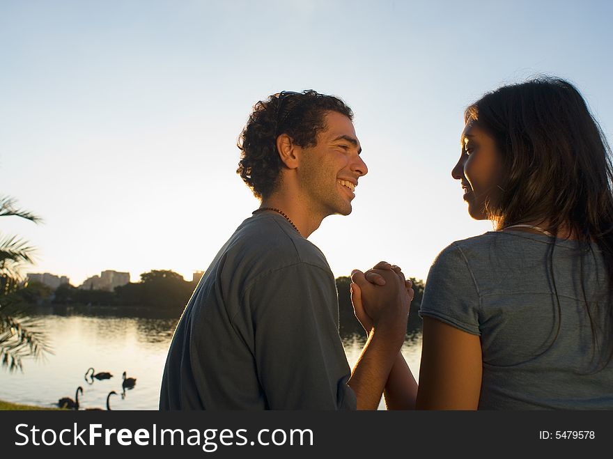 Happy couple clasp hands as they smile at each other. There is a pond with swans and buildings in the background. Horizontally framed photograph. Happy couple clasp hands as they smile at each other. There is a pond with swans and buildings in the background. Horizontally framed photograph