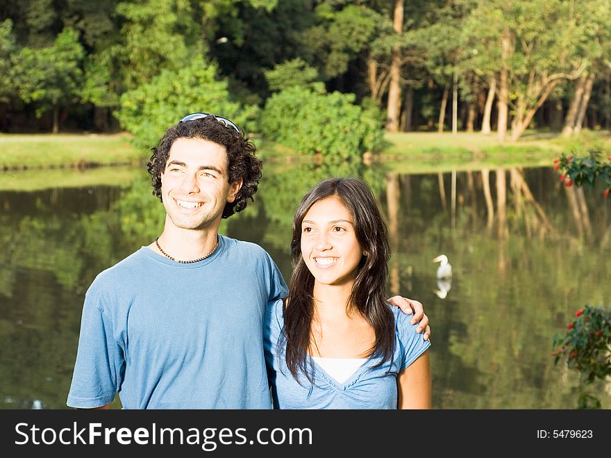 Happy couple smiling in front of a pond. He has his arm around her as they stare off in the distance. Horizontally framed photograph. Happy couple smiling in front of a pond. He has his arm around her as they stare off in the distance. Horizontally framed photograph