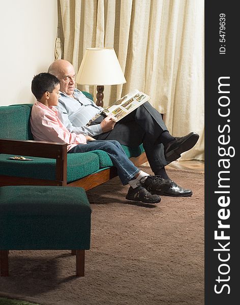 Grandfather and grandson sit on couch reading a book. Grandfather points to a picture on the page as grandson watches. This is a vertically framed photo. Grandfather and grandson sit on couch reading a book. Grandfather points to a picture on the page as grandson watches. This is a vertically framed photo.