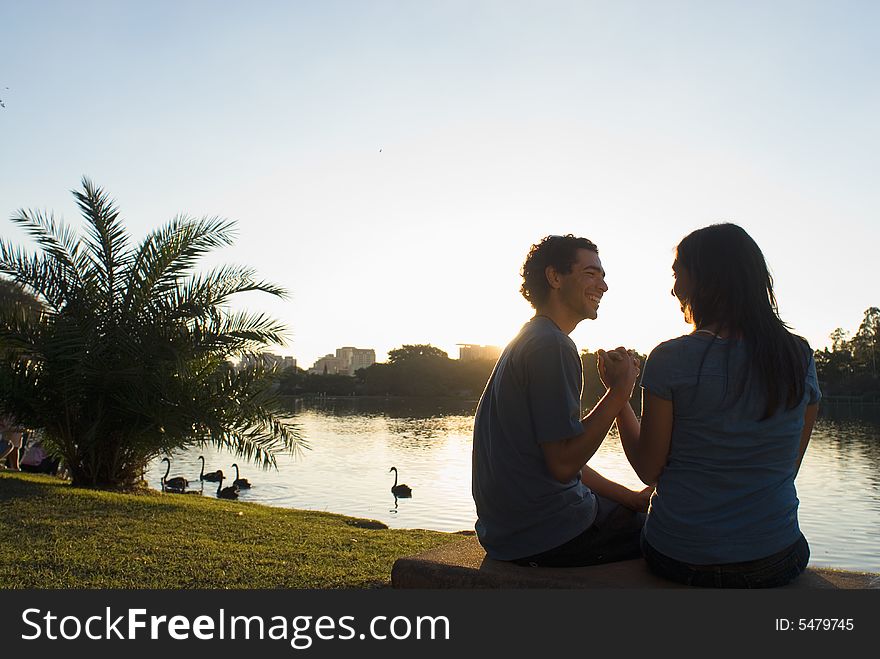 Happy couple clasp hands as they smile at each other. There is a pond with swans and buildings in the background. Horizontally framed photograph. Happy couple clasp hands as they smile at each other. There is a pond with swans and buildings in the background. Horizontally framed photograph