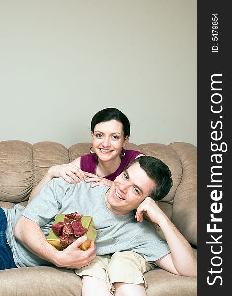 A smiling man holding a gift box lying on a smiling woman's lap on sofa. Vertically framed shot. A smiling man holding a gift box lying on a smiling woman's lap on sofa. Vertically framed shot.