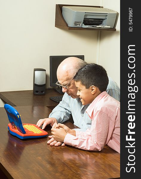 Grandfather and Grandson are seated at a table looking at a blue toy computer. There is a computer and a printer in the background. Vertically framed photograph. Grandfather and Grandson are seated at a table looking at a blue toy computer. There is a computer and a printer in the background. Vertically framed photograph.