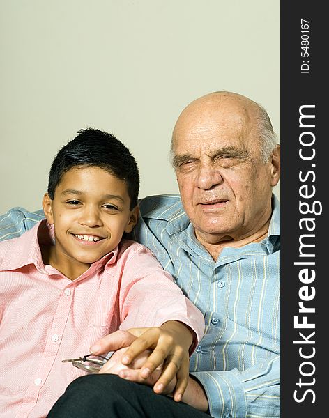 Grandfather and grandson are sitting on the couch looking forward happily. They hold each others hand with happy expressions. This is a vertically framed photo. Grandfather and grandson are sitting on the couch looking forward happily. They hold each others hand with happy expressions. This is a vertically framed photo.