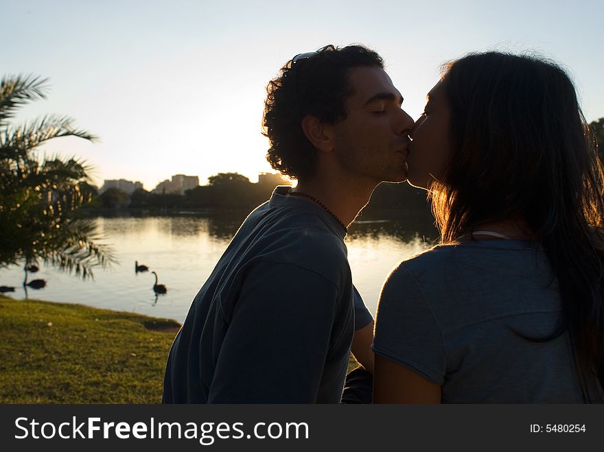 Couple kissing. There is a pond with swans, and buildings in the background. Horizontally framed photograph. Couple kissing. There is a pond with swans, and buildings in the background. Horizontally framed photograph