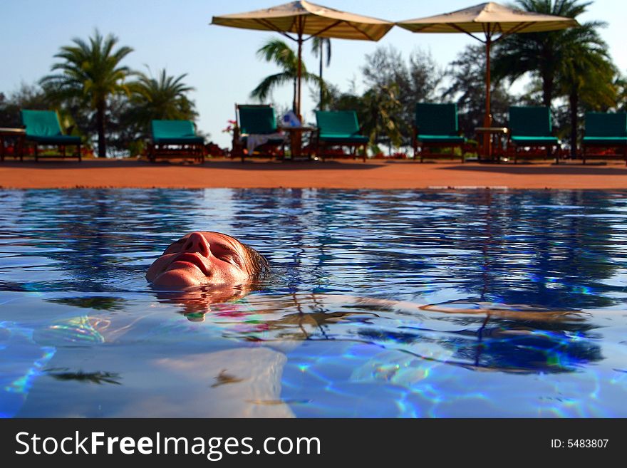 A young woman is relaxing in the pool. Ideal vacation shot with sun umbrellas and palm trees in the back.