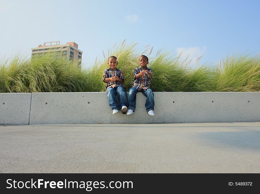 Brothers sitting on a park ledge with greenery in the background. Brothers sitting on a park ledge with greenery in the background.
