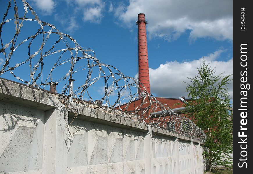 A secured industrial zone with concrete fence, barbed wire and brick chimney-stalk on the blue sky background.