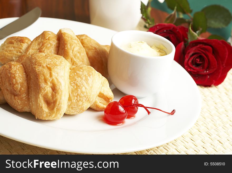 Breakfast plate with croissants, butter and glazed maraschino cherries with red roses in the background. Breakfast plate with croissants, butter and glazed maraschino cherries with red roses in the background