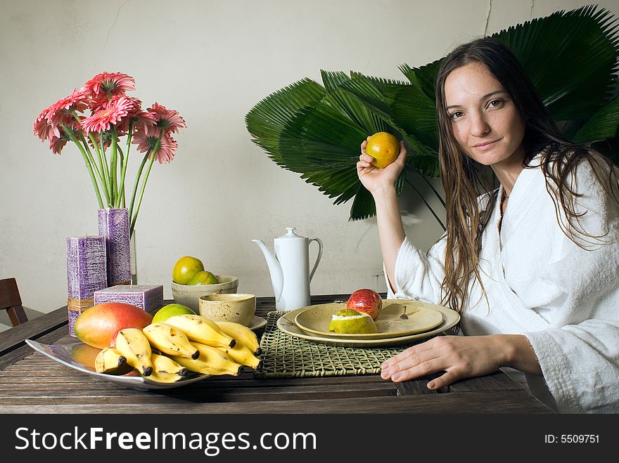 Woman smiles as she has breakfast. She is holding an orange. There are many fruits on the table. Horizontally framed photograph. Woman smiles as she has breakfast. She is holding an orange. There are many fruits on the table. Horizontally framed photograph.