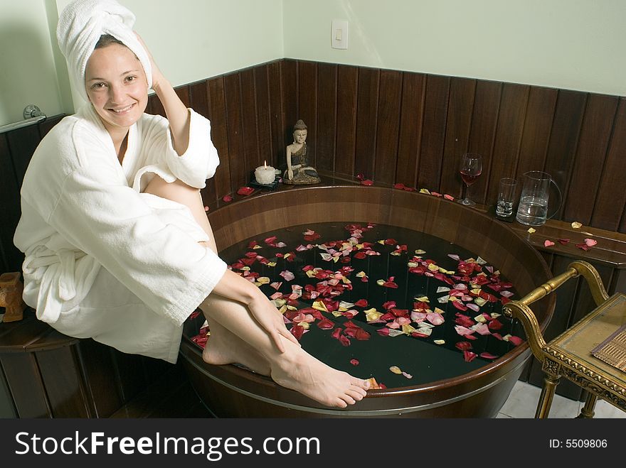 Woman in bathrobe relaxes by a spa tub filled with rose petals as she smiles. Horizontally framed photograph. Woman in bathrobe relaxes by a spa tub filled with rose petals as she smiles. Horizontally framed photograph