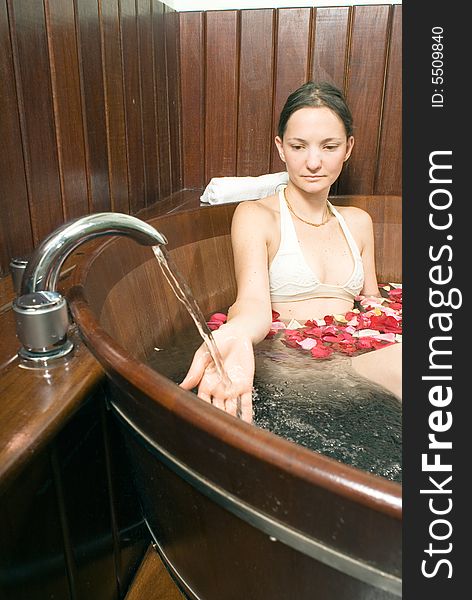 Young attractive lady, relaxing in a bathtub, playing with the running faucet. Multicolored rose petals float on the surface of the bathtub. Vertically framed shot. Young attractive lady, relaxing in a bathtub, playing with the running faucet. Multicolored rose petals float on the surface of the bathtub. Vertically framed shot.