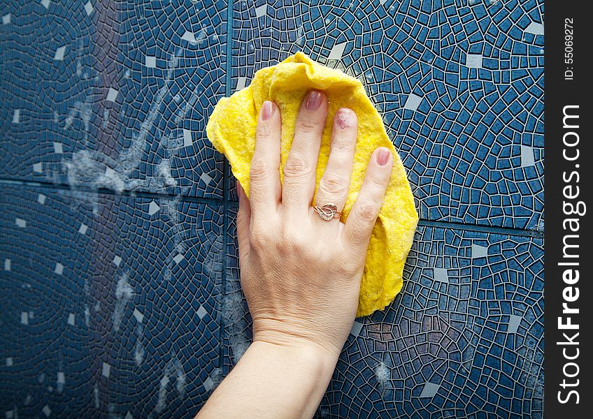 Female hand washes the blue tile on the wall with a yellow cloth lather in the bathroom closeup. Female hand washes the blue tile on the wall with a yellow cloth lather in the bathroom closeup