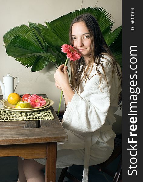 Woman smells a pink daisy as she is having fruit for breakfast. There is a large plant behind her. Vertically framed photograph. Woman smells a pink daisy as she is having fruit for breakfast. There is a large plant behind her. Vertically framed photograph.