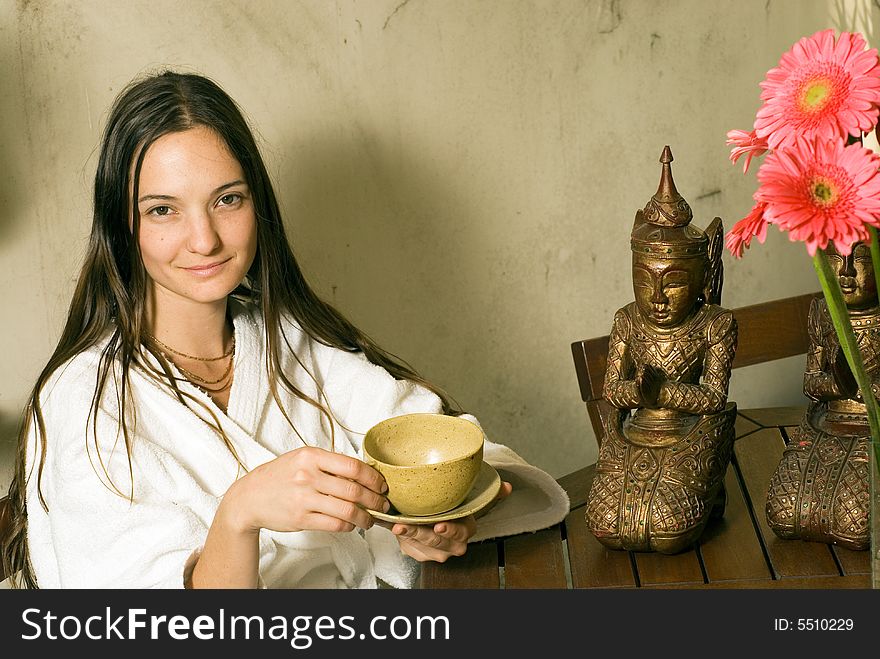 Woman smiles as she holds a tea cup. She sits next to a bronze statue and pink flowers. Horizontally framed photograph. Woman smiles as she holds a tea cup. She sits next to a bronze statue and pink flowers. Horizontally framed photograph.