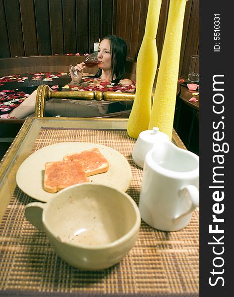 Woman relaxes in a spa tub filled with rose petals. She is drinking red wine. There are candles and lunch in the foreground. Woman relaxes in a spa tub filled with rose petals. She is drinking red wine. There are candles and lunch in the foreground.