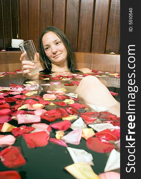 Woman smiles she looks at a glass of water. She is relaxing in a spa tub filled with rose petals. Vertically framed photograph. Woman smiles she looks at a glass of water. She is relaxing in a spa tub filled with rose petals. Vertically framed photograph.