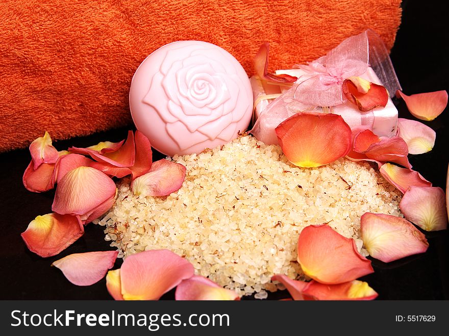 Bath salt and soap with a pink ribbon, with rose petals and bright orange towel laying on black granite. Bath salt and soap with a pink ribbon, with rose petals and bright orange towel laying on black granite