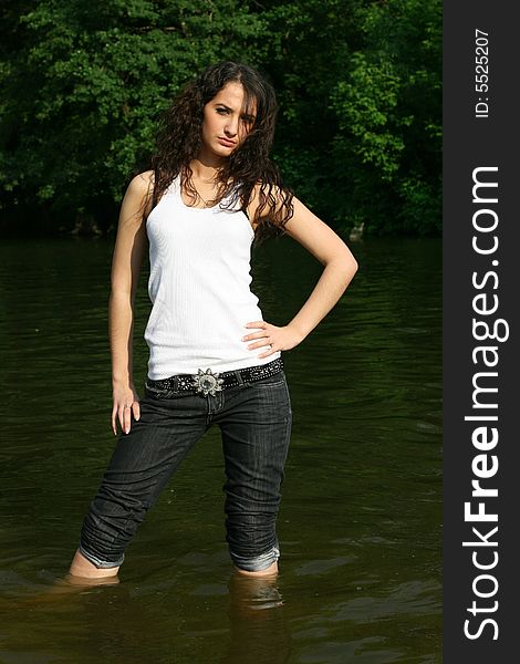 The girl in dark blue jeans and a white vest costs in water on a background of green trees. The girl in dark blue jeans and a white vest costs in water on a background of green trees