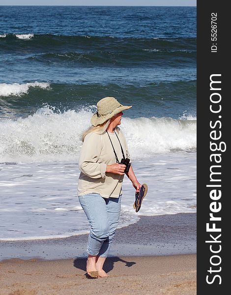 Woman wearing straw hat carrying binoculars and sandals walking on beach. Waves splashing in background. Woman wearing straw hat carrying binoculars and sandals walking on beach. Waves splashing in background.