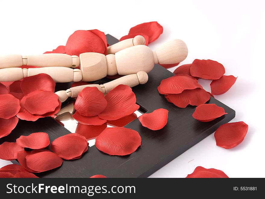 A wooden man submerged in red rose petal, lying on a black wide border mirror. A wooden man submerged in red rose petal, lying on a black wide border mirror.