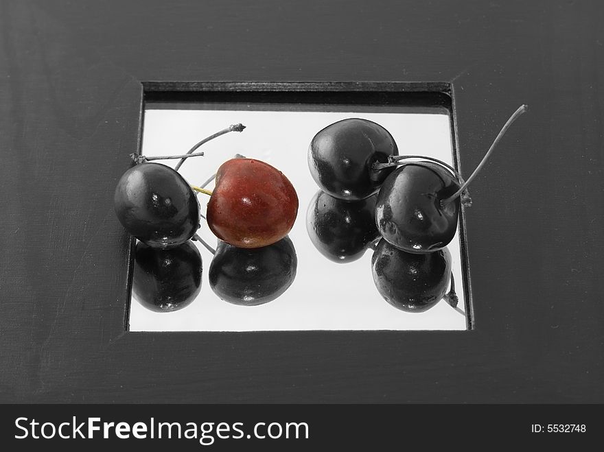 A red cherry surrounded by three black cherries on a mirror with wide black border.